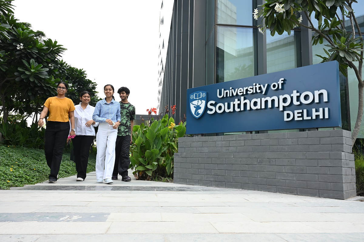A group of smiling students walk towards the camera past a large University of Southampton Delhi logo